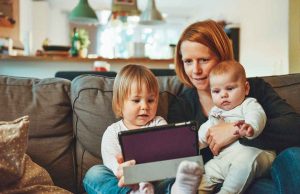 woman with red hair with two babies in white holding ipad on brown couch