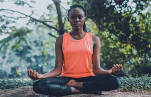 black woman in orange shirt and black leggings meditating outside in wooded area