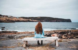 woman with red hair in teal jacket on bench by water