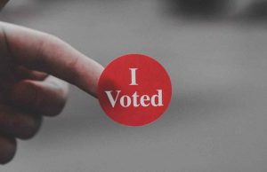grey background with hand holding a round red sticker that says i voted