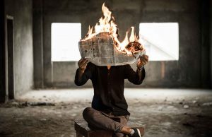 person in black shirt and grey pants sitting in grey room with newspaper on fire