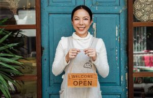 woman in front of teal door holding open sign
